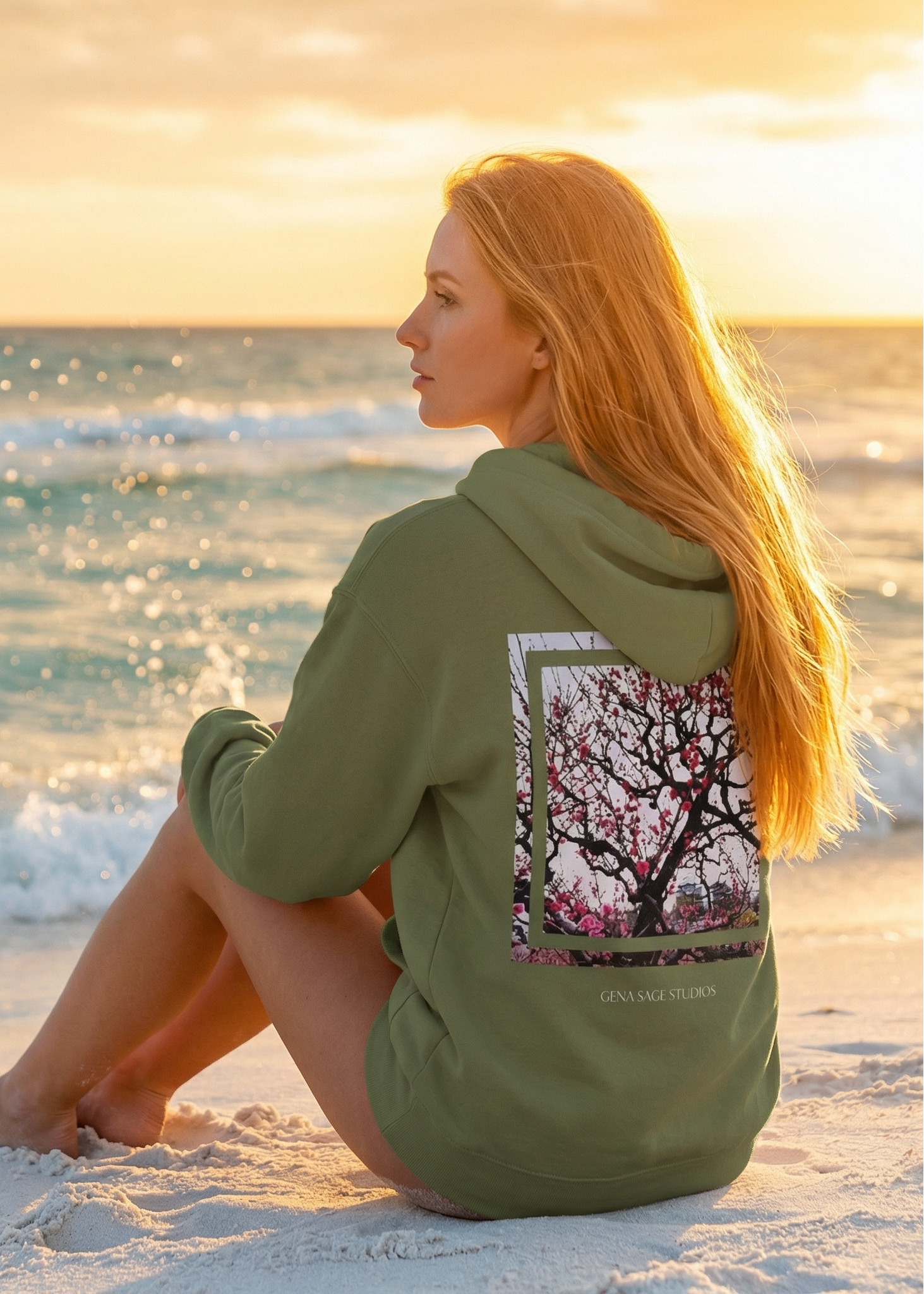Woman sitting on a beach wearing a green hoodie with a tree design, looking at the ocean.