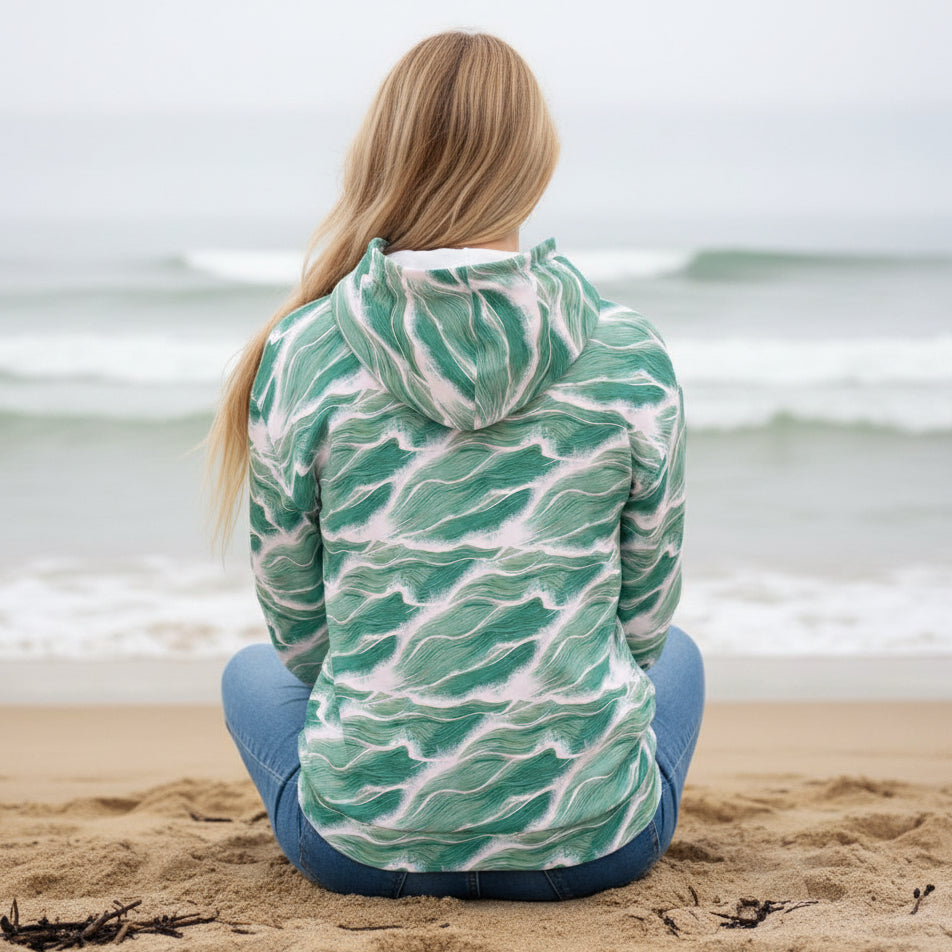 Back view of Sage Seafoam Wave Hoodie by Chameleon Surf featuring flowing sage green and white wave pattern sitting on the beach facing the ocean