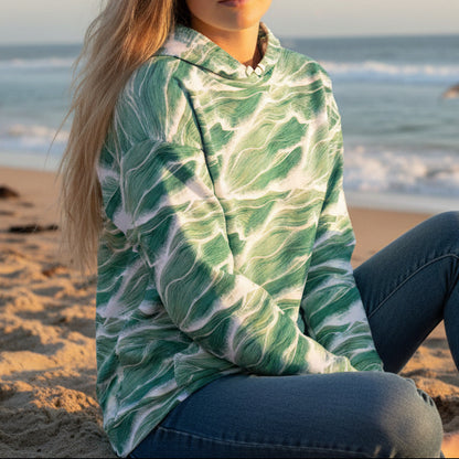 Model wearing Sage Seafoam Wave Hoodie by Chameleon Surf featuring flowing sage green and white wave pattern sitting on the beach